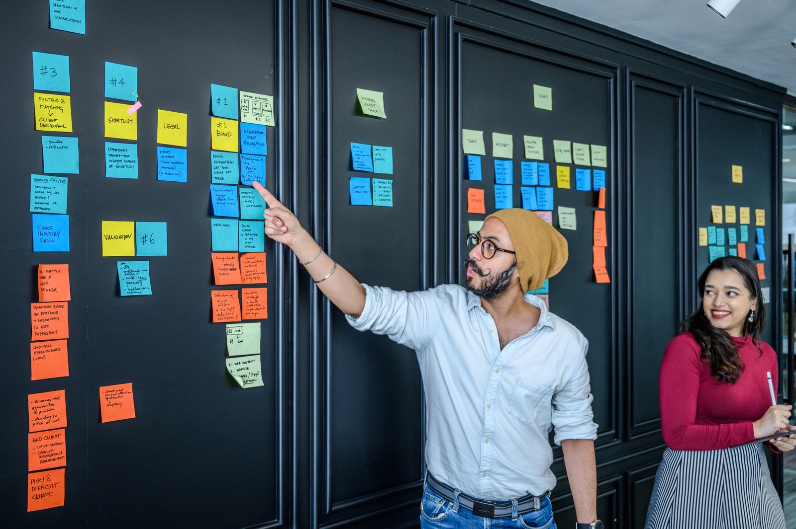 Man pointing at coloured sticky notes on a board while planning a project with a colleague